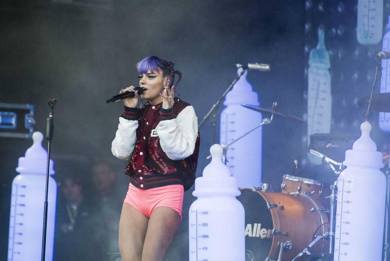 Lily Allen performing on stage with purple hair, a red sequined varsity jacket, and pink shorts, flanked by giant prop baby bottles.