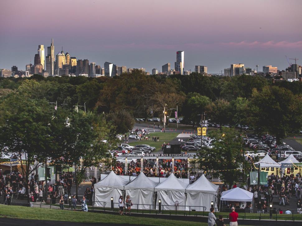 Dusk over the Mann Center in Fairmount Park, white event tents and a crowd gathered with the Philadelphia skyline behind them.