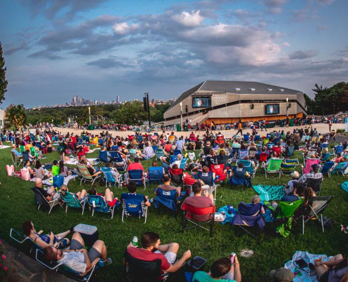 A large crowd relaxing in lawn chairs on the grass at the Mann Center at sunset, the wooden pavilion and Philadelphia skyline in the distance.